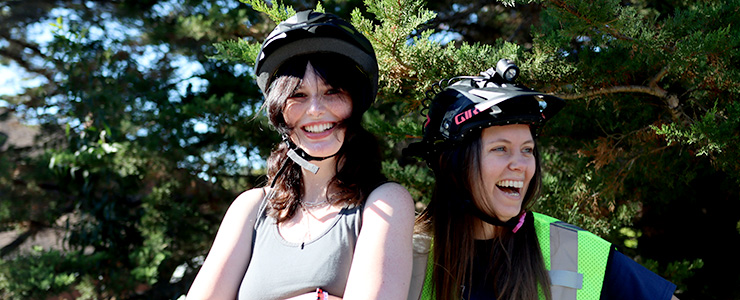 Two young women standing in front of greenery while smiling and wearing bike helmets
